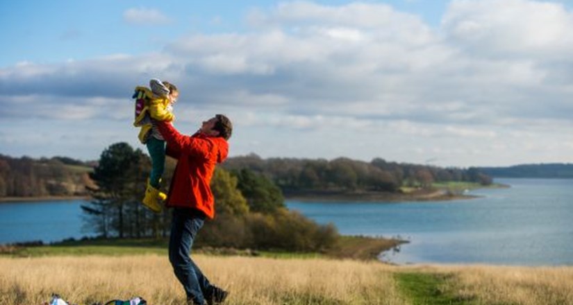 Father lifting child near a lake on a sunny day.