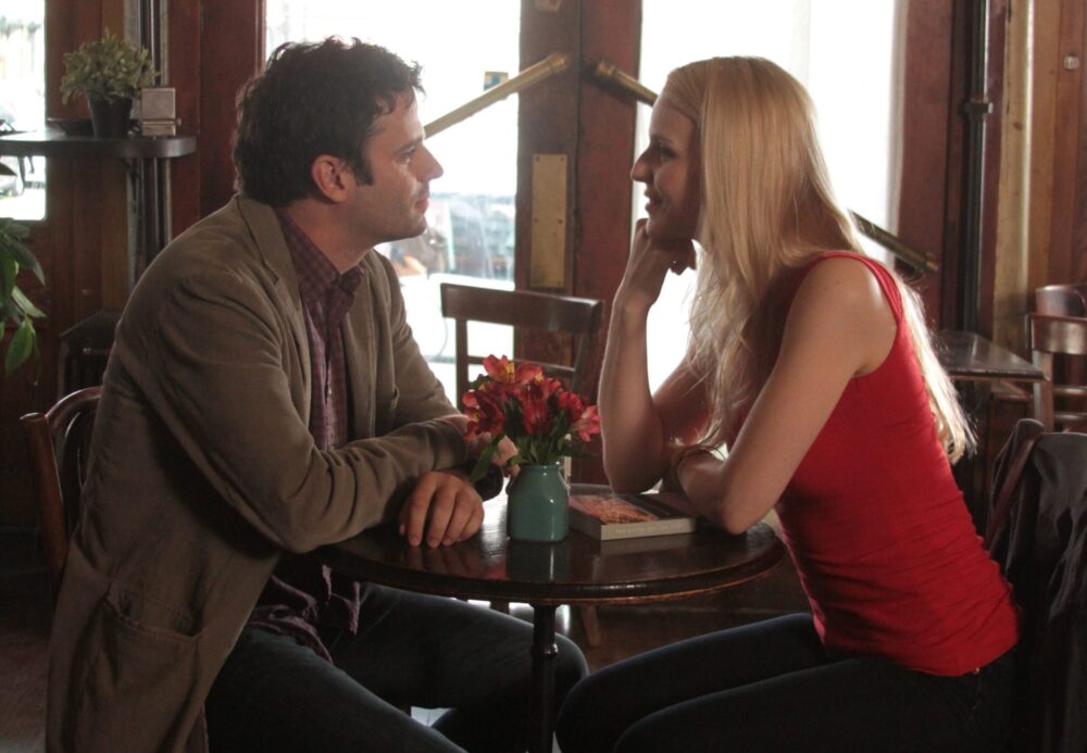 Couple seated at cafe table with flowers, engaged in conversation.