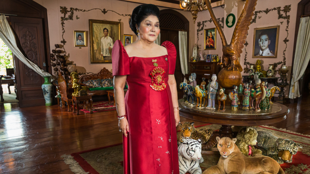 Imelda Marcos in red dress, surrounded by opulent decor.