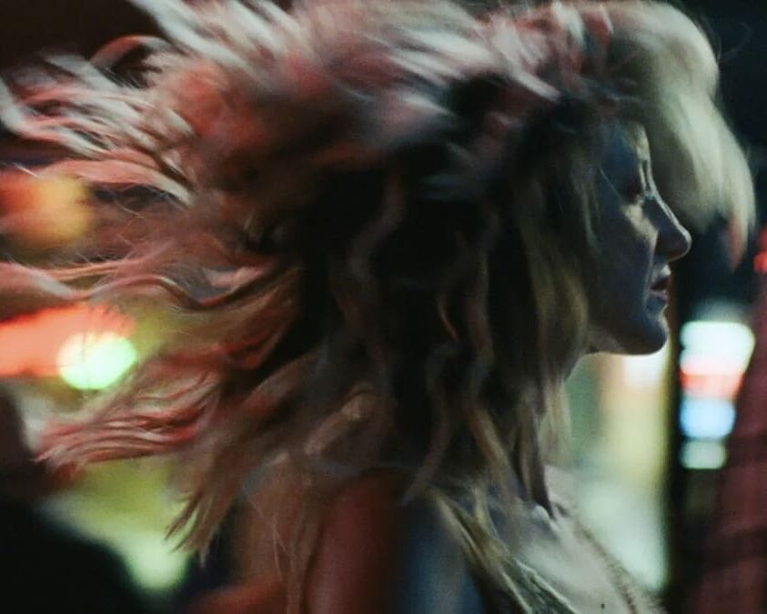 To Leslie: Woman with windswept blonde hair, side profile, in a dimly lit setting.
