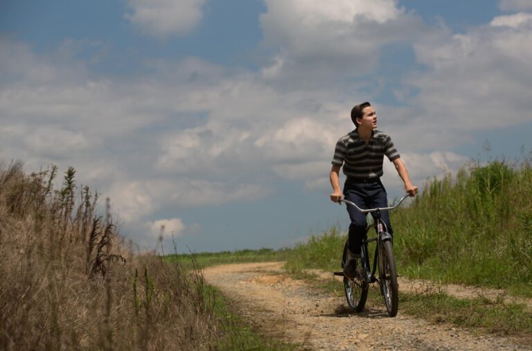 Boy riding a bicycle on a dirt road in "Ithaca," looking off to the side.