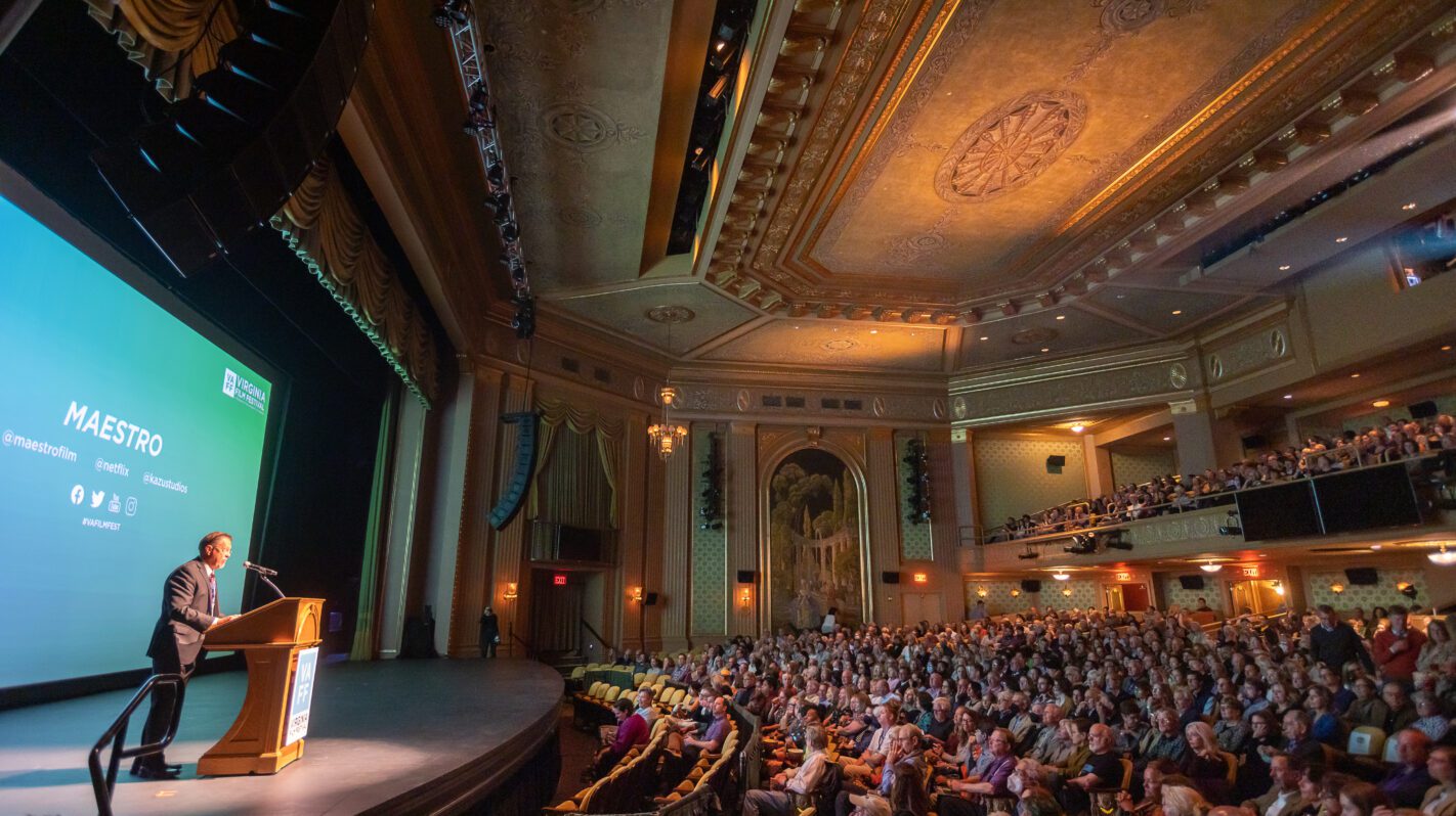 Speaker on stage at Virginia Film Festival, Maestro film presentation. Large audience in ornate theater.