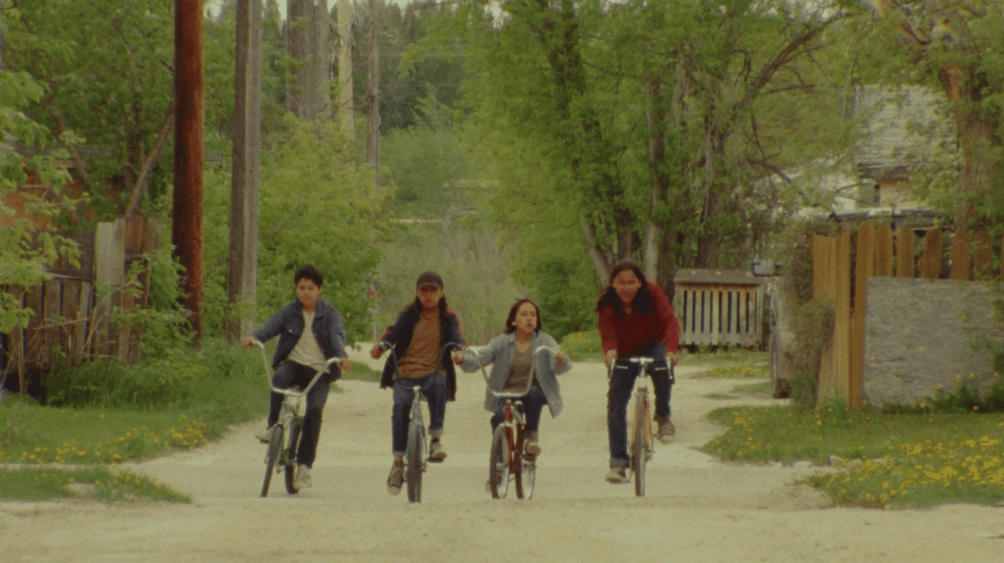 Four Indigenous children ride bikes down a dirt road, embodying a sense of community and childhood.