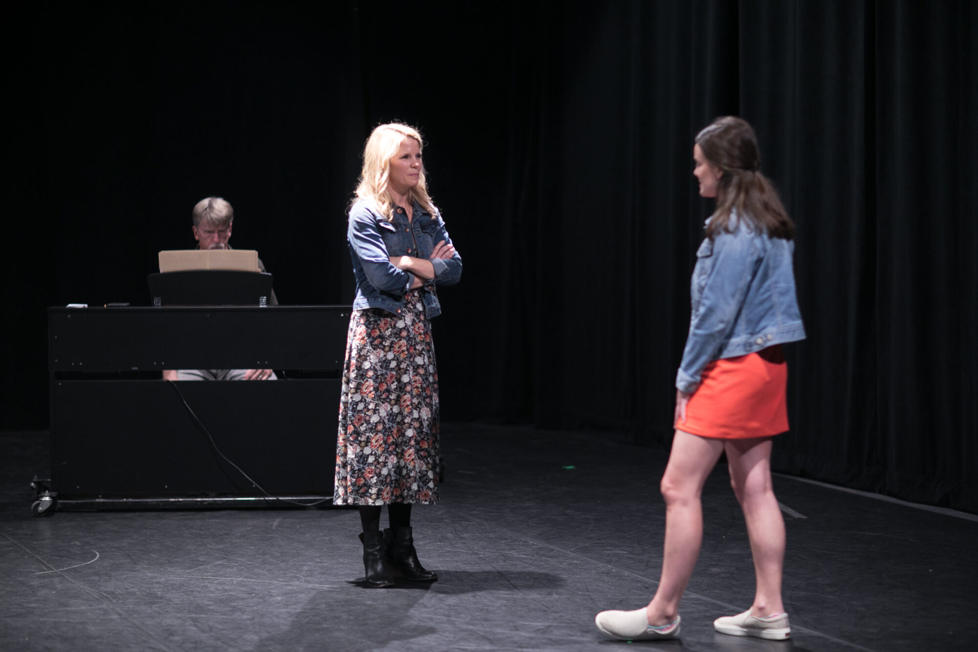Two women in a stage scene, with a man at a piano in the background.