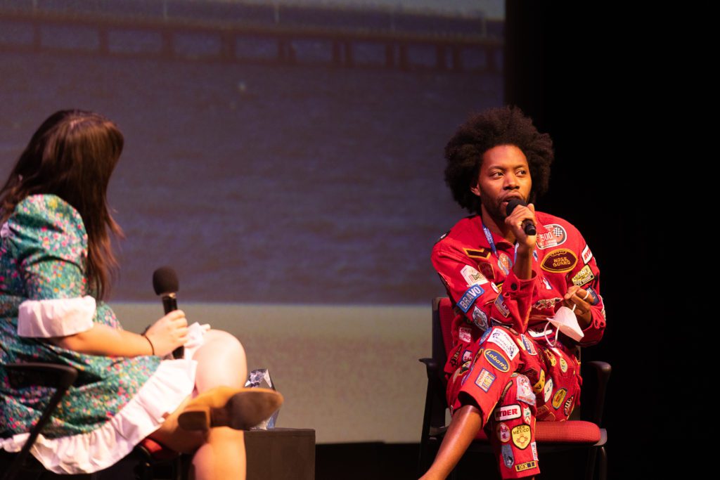 Man speaking at NEA 2023 event, wearing a red jumpsuit.