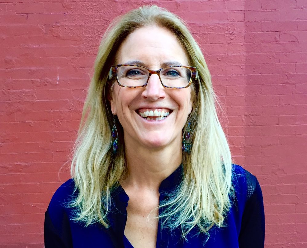 Smiling woman with glasses and long blonde hair stands against a red brick wall.