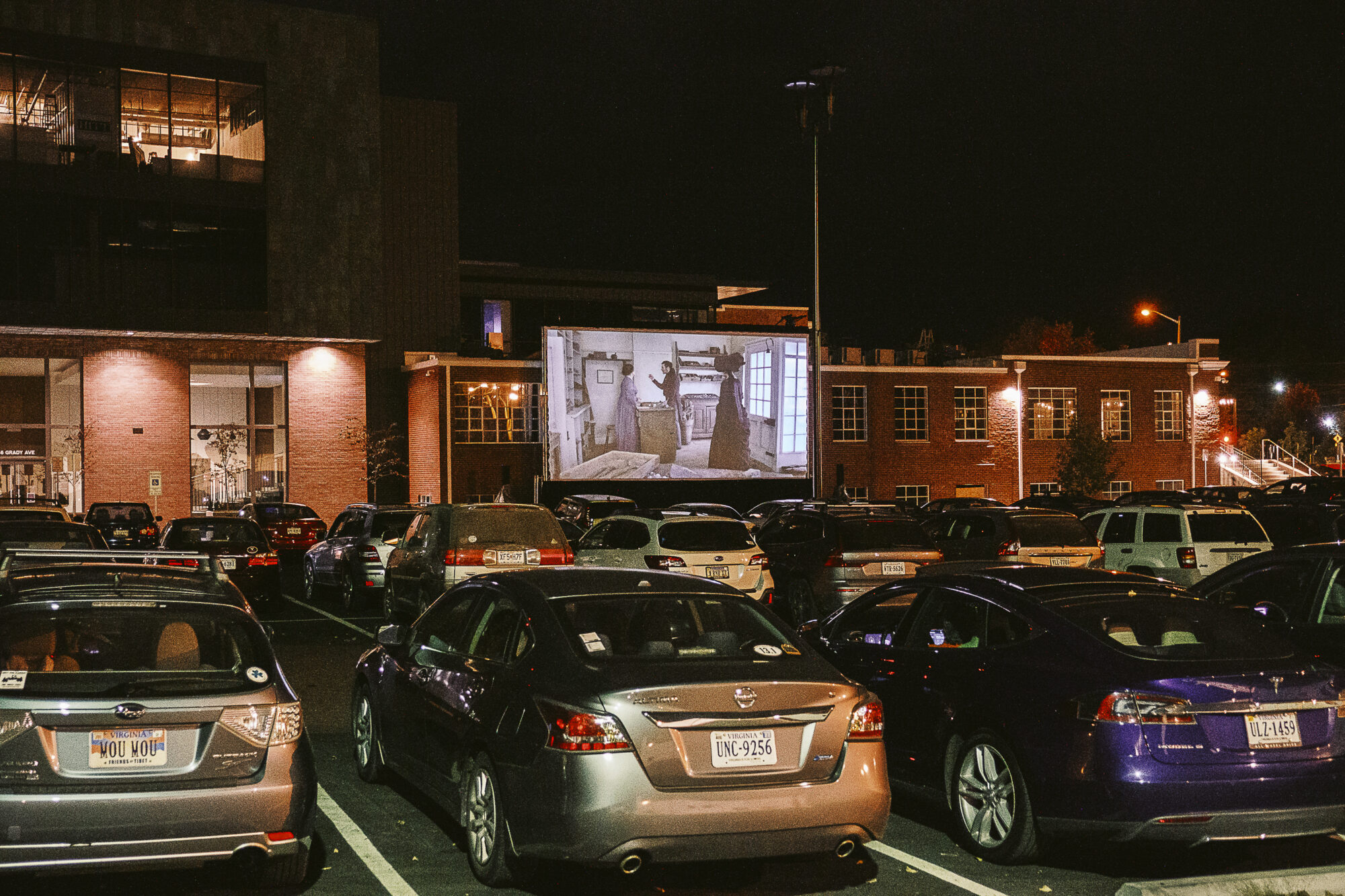 Cars parked at a drive-in movie theater showing a film on a large outdoor screen at night.
