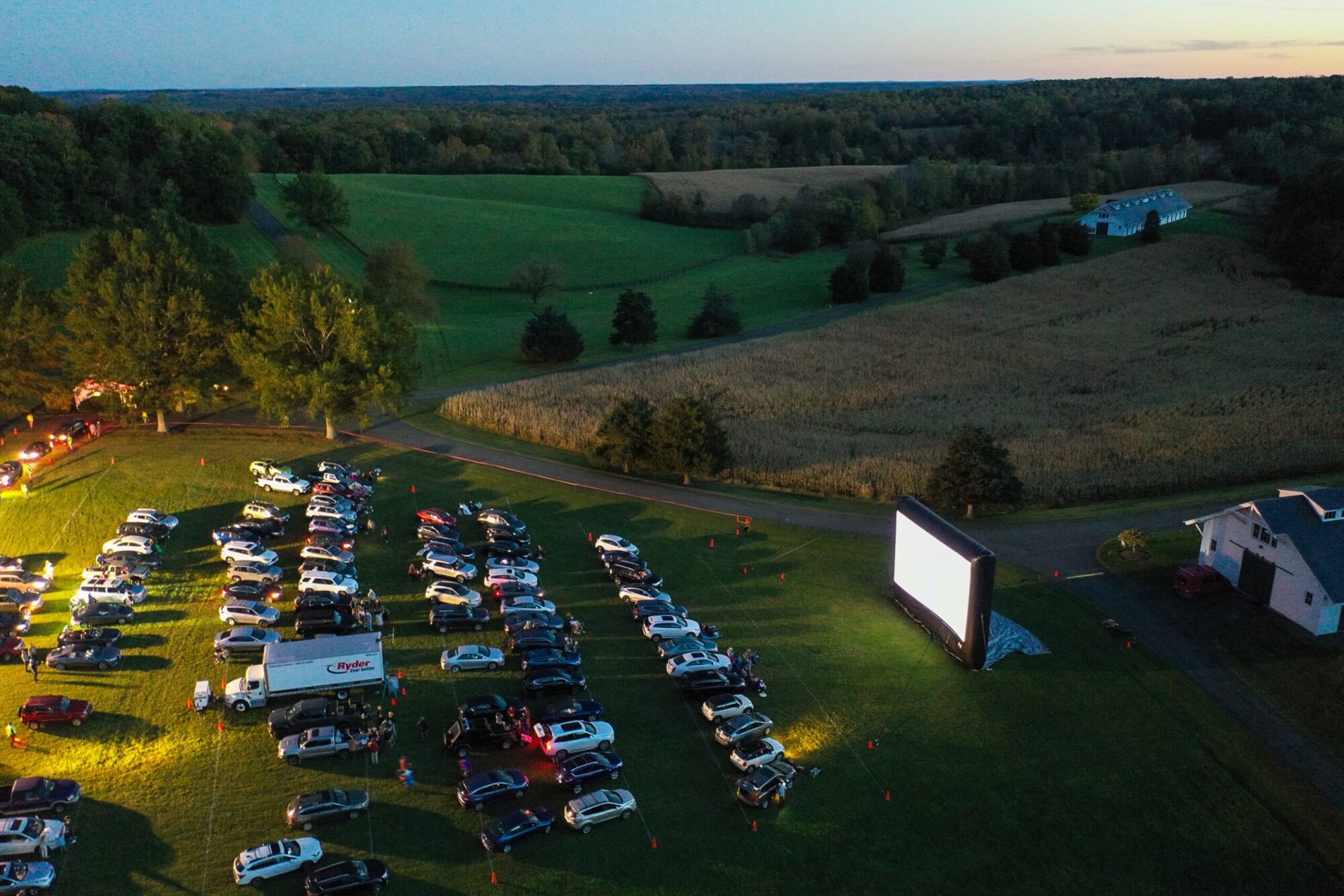 Drive-in movie night on a farm field, cars parked facing the screen. Ryder truck visible.