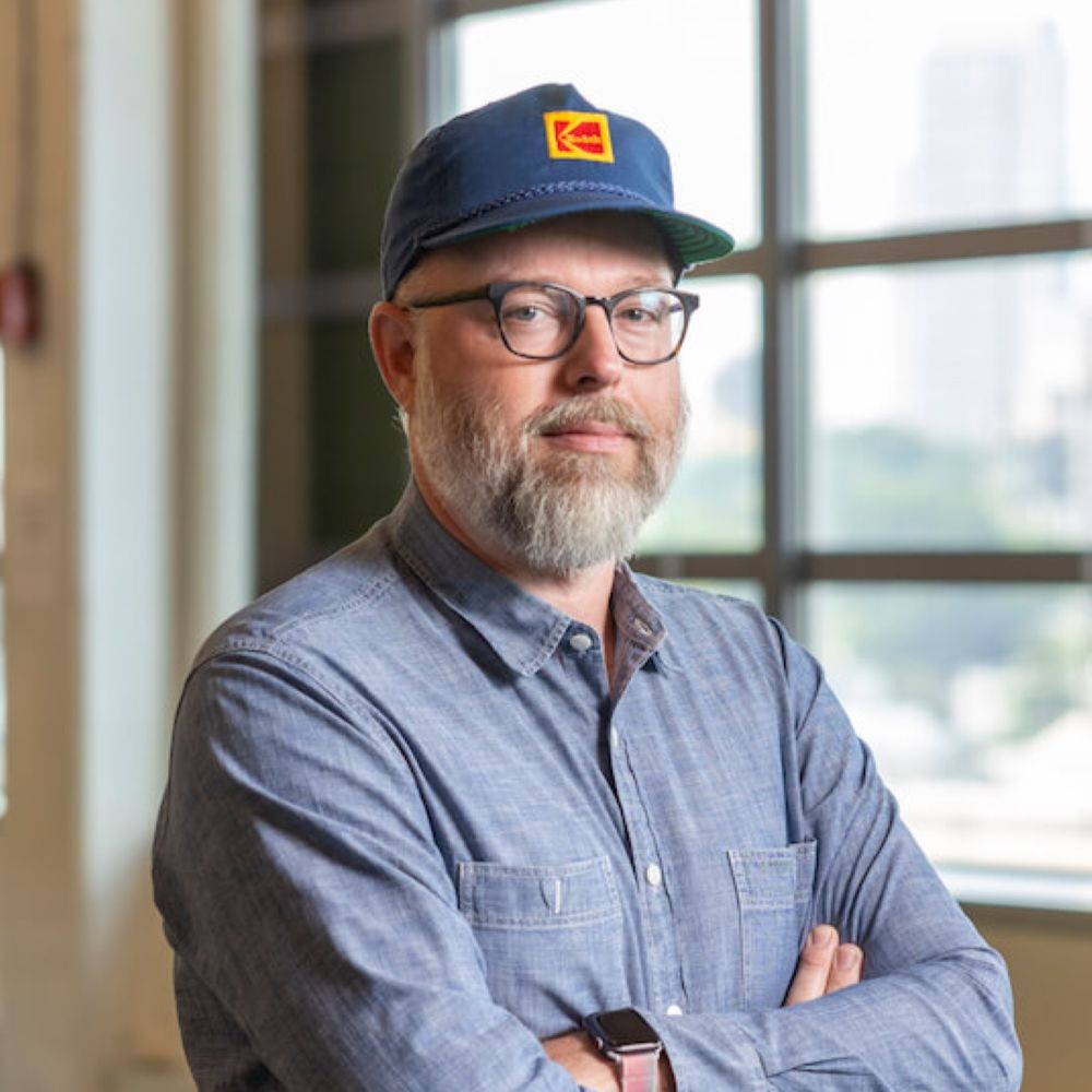 Charles Haine wearing a Kodak hat, glasses, and a blue shirt, arms crossed.