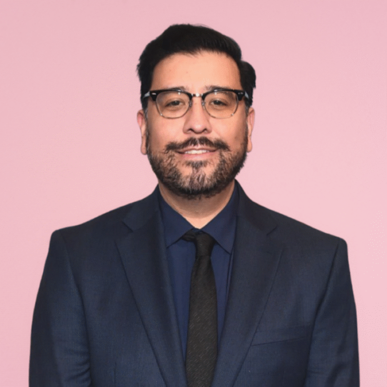 Ernie Bustamante in a suit and tie, smiling against a pink background.