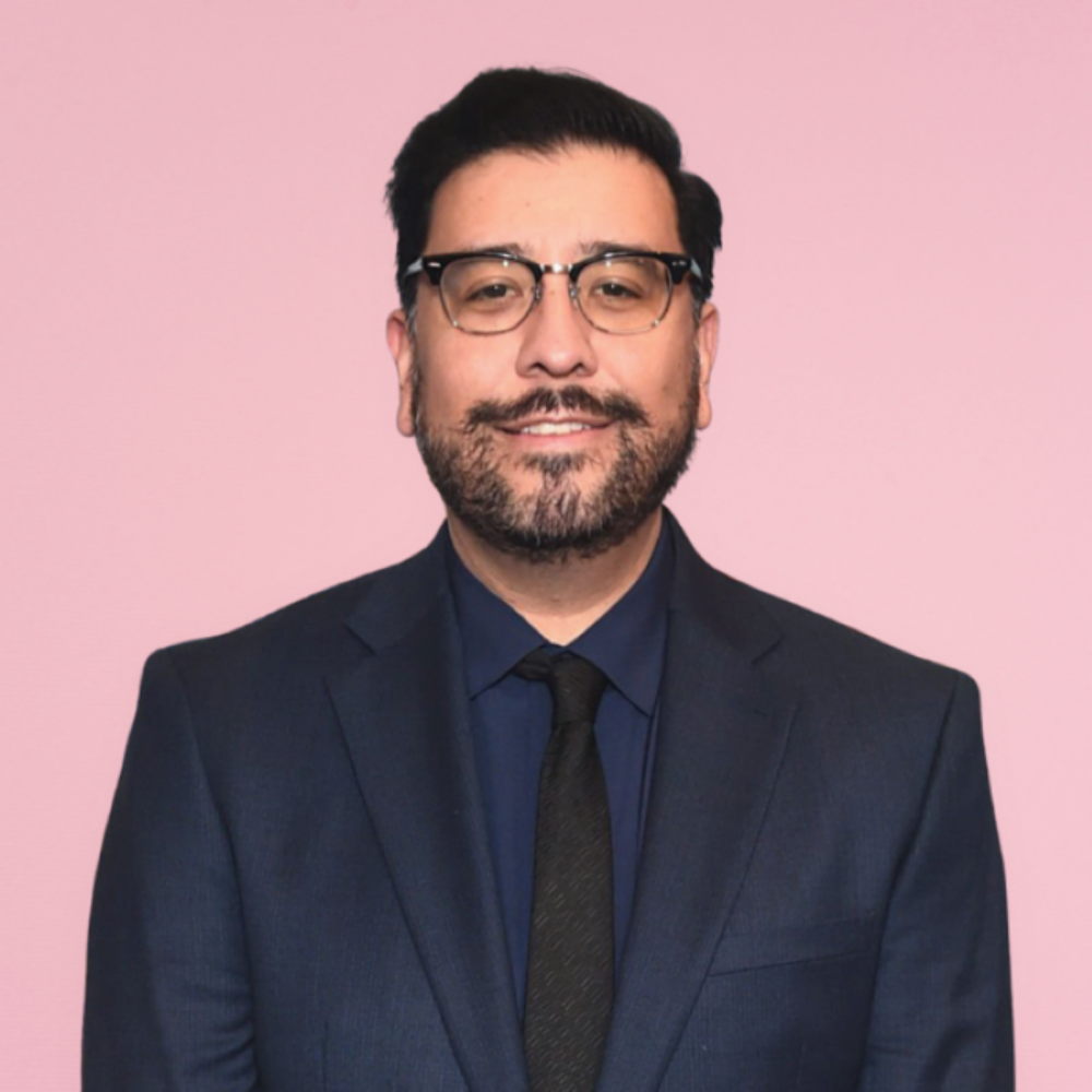 Ernie Bustamante in a suit and tie, smiling against a pink background.