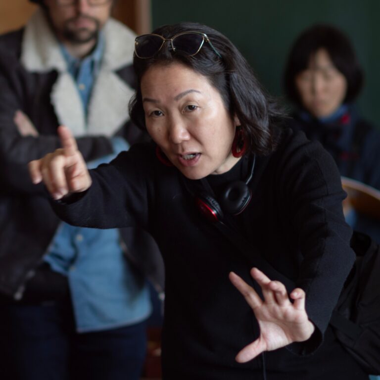 Director HIKARI gesturing while filming a scene, wearing headphones and sunglasses on her head.