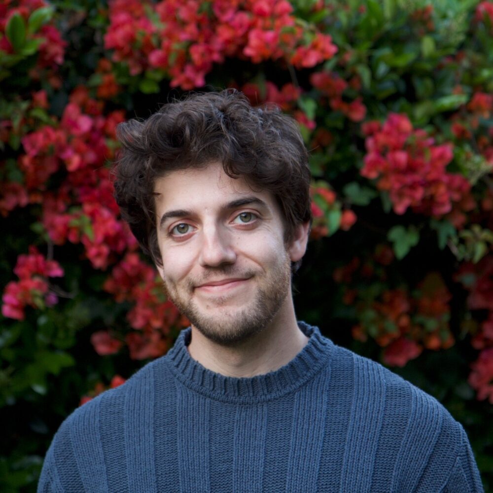 Jacob Seltzer smiling in front of vibrant red bougainvillea flowers.