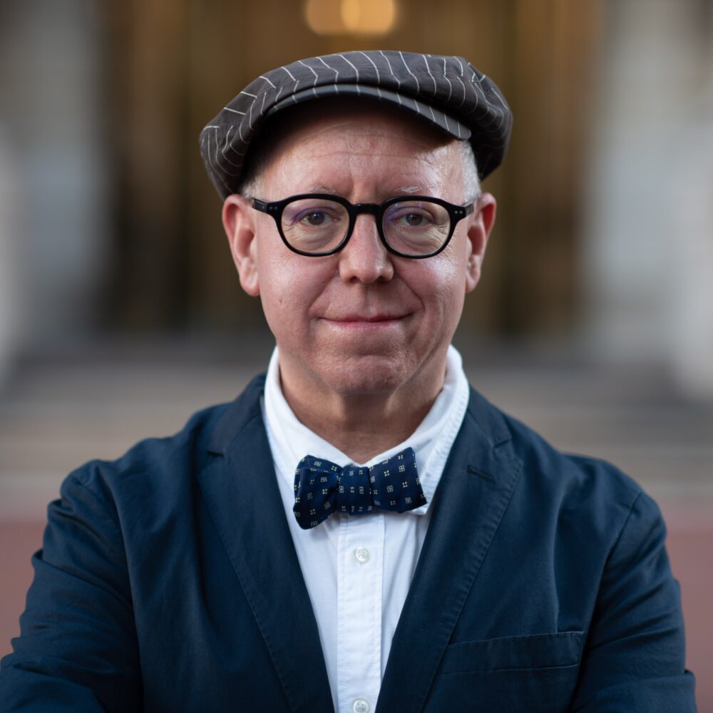 James Schamus wearing a newsboy cap, bow tie, and glasses, smiling at the camera.