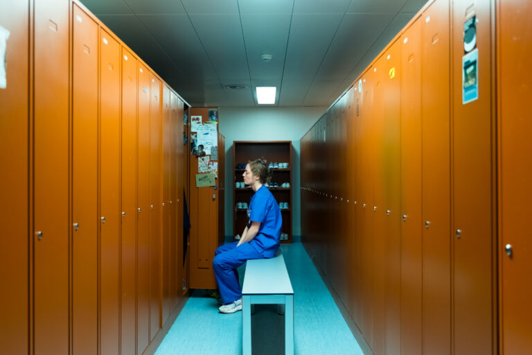 Healthcare worker in blue scrubs sits in locker room.