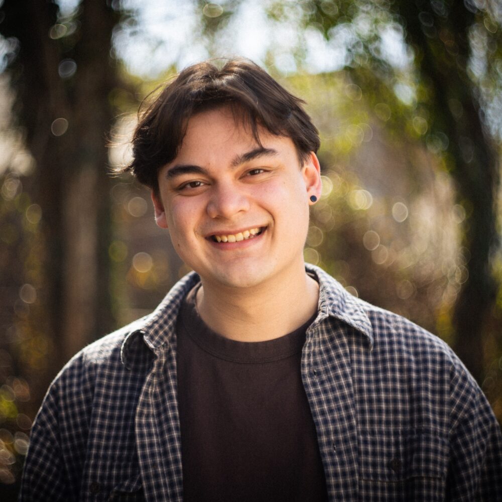 Luca Huff smiling outdoors wearing a flannel shirt and dark t-shirt.