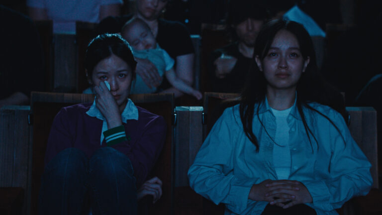 Two women with tears in their eyes sit in a dark movie theater, evoking nervous energy.