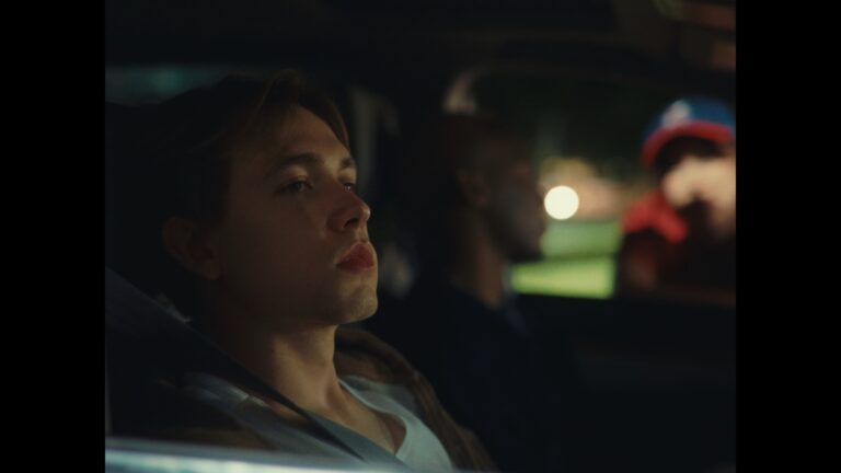 Pensive young man in car at night. Group of friends visible in background.