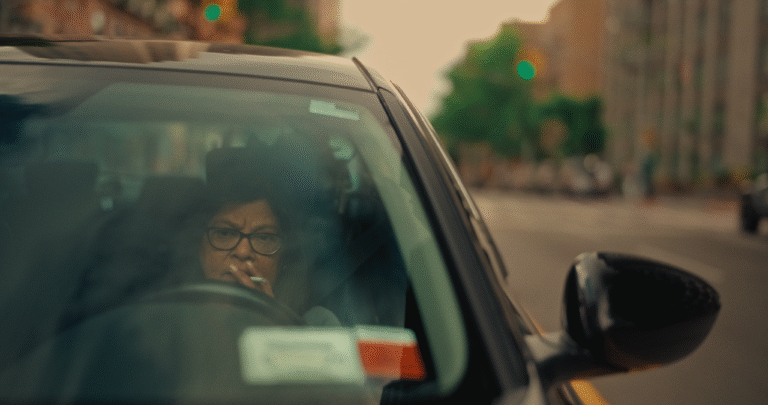 Woman with glasses smoking in car, city street visible through windshield. "Shanti Rides Shotgun