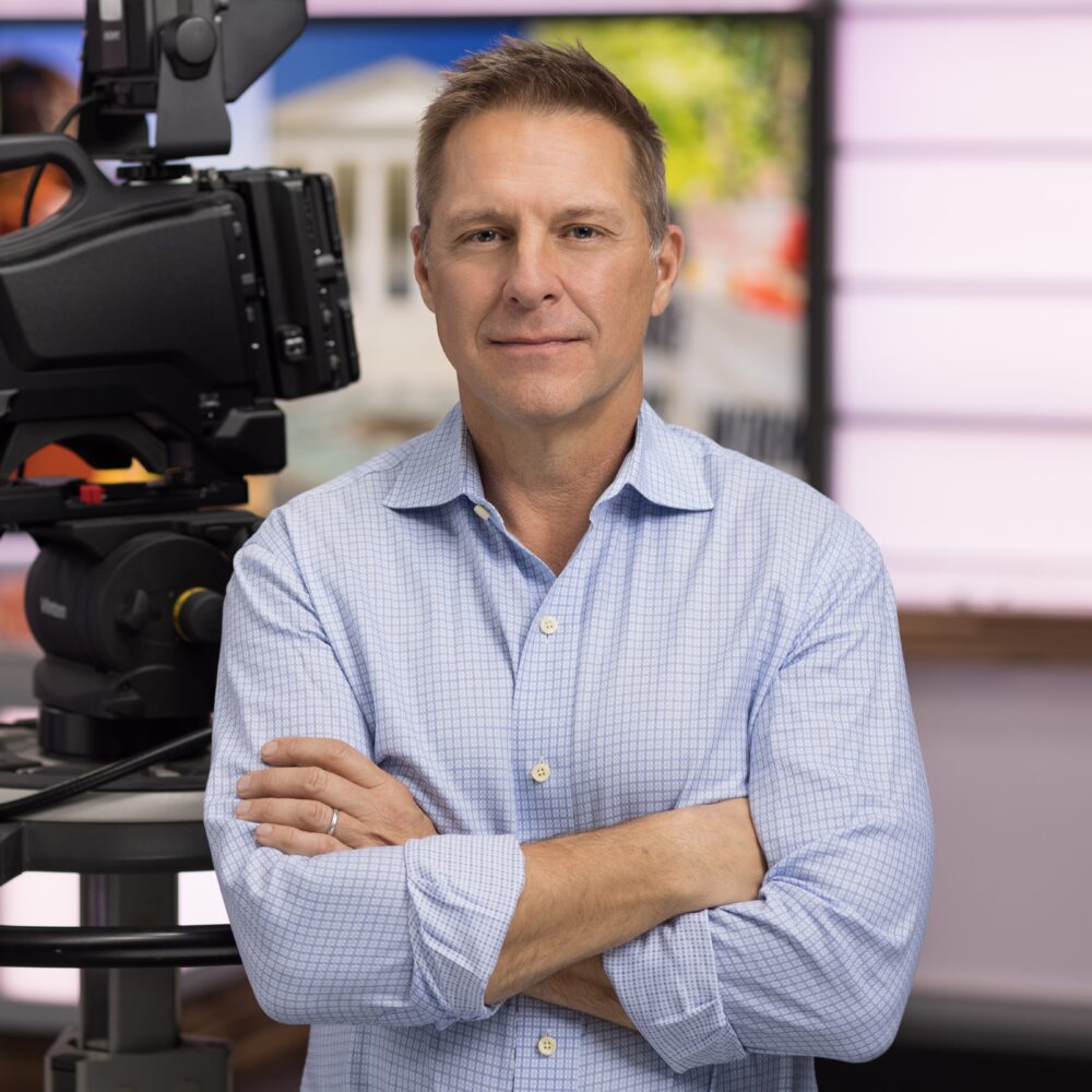 Steve Humble poses with arms crossed next to a professional video camera in a studio setting.