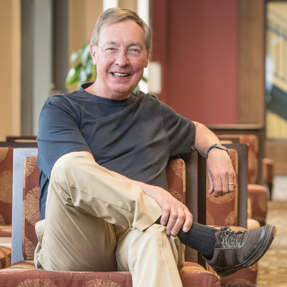 Ted Dintersmith sits smiling in a chair, wearing khaki pants and a black shirt.