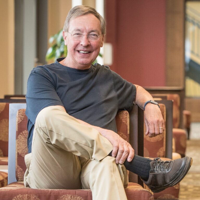 Ted Dintersmith sits smiling in a chair, wearing khaki pants and a black shirt.