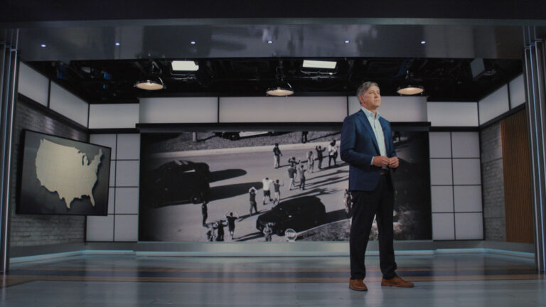 Man in a suit stands in a studio with a US map and a black and white photo.