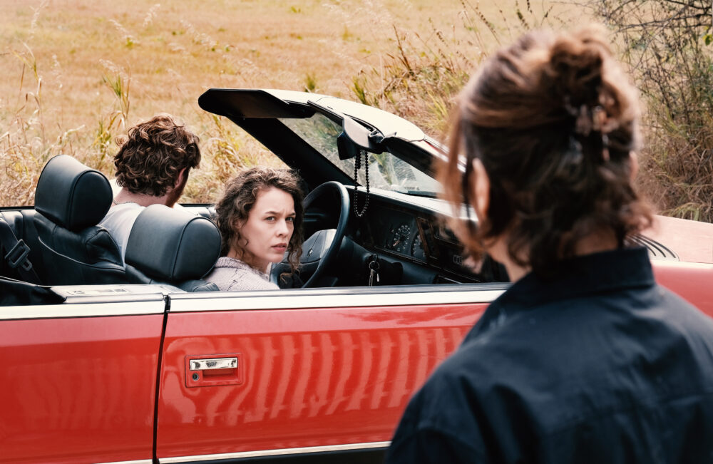 Two people in a red convertible, woman in front looking back, man in back seat