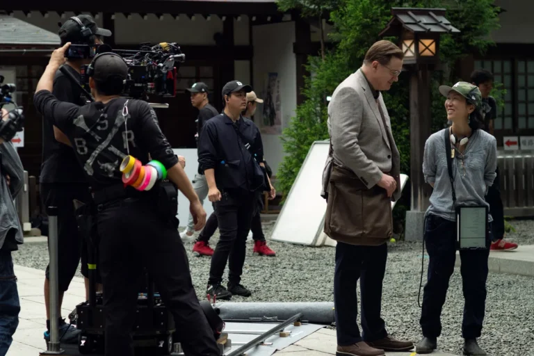 On set of 'Rental Family' film: Actor in suit speaking with crew member outdoors.