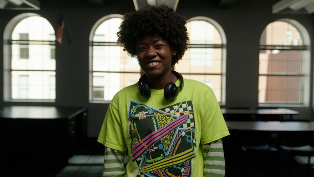Film still from IMMUTABLE. Smiling young person with afro, headphones, and bright shirt in library setting.